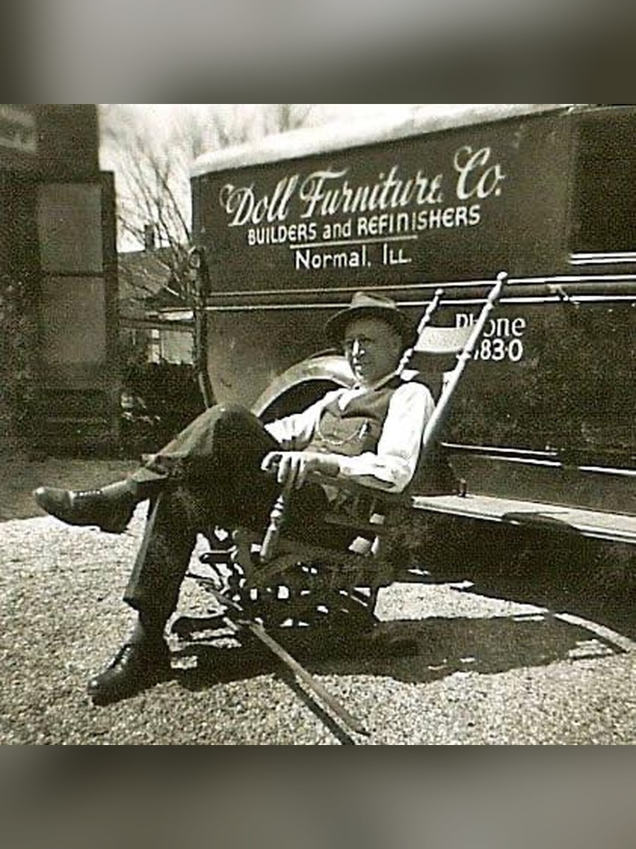 A.E. Shutt, founder of Doll Furniture Company, seated in a rocking chair in front of the original delivery truck reading "Doll Furniture Co. Builders and Refinishers, Normal, Ill."