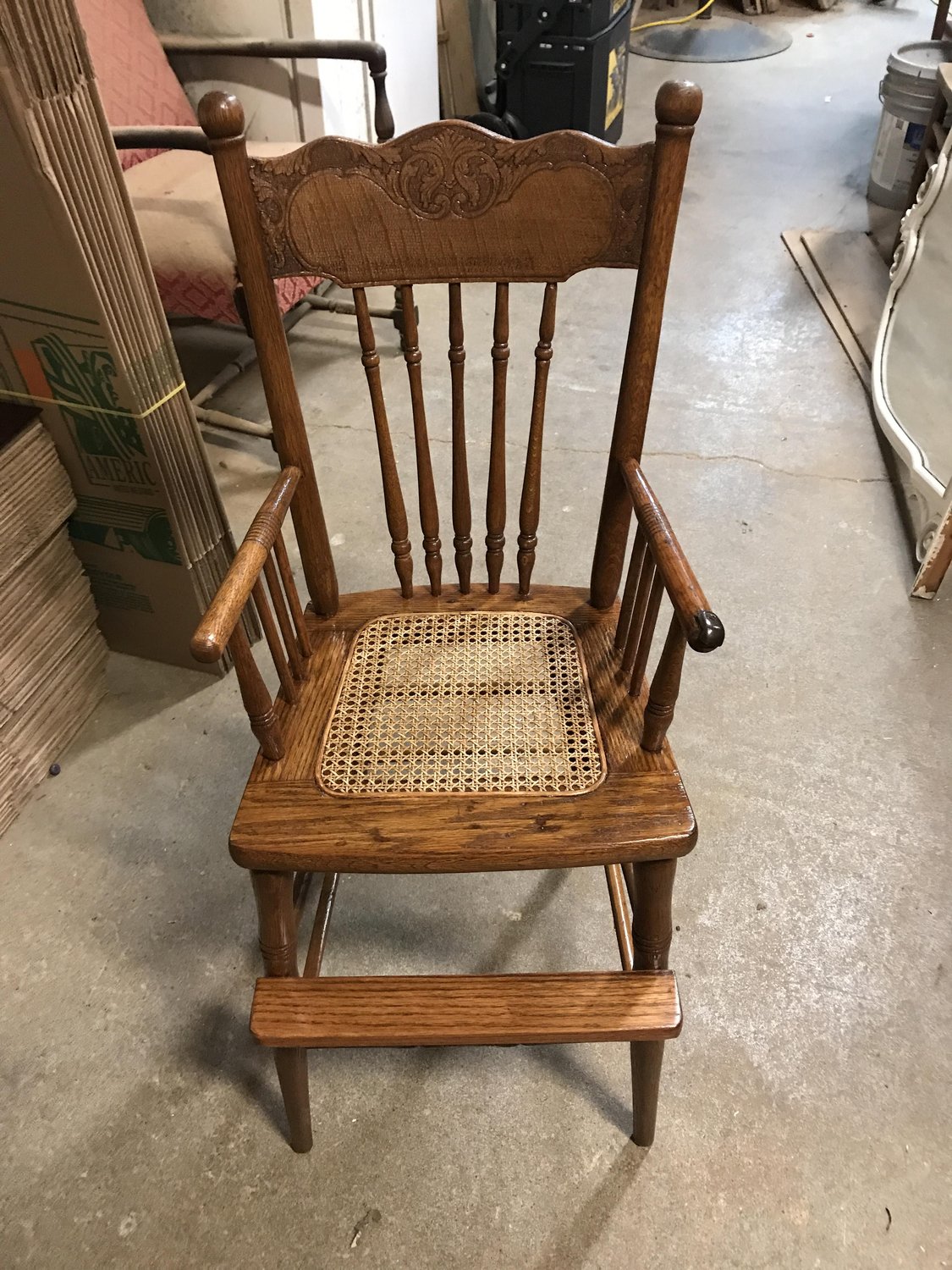Antique press-back oak youth chair with carved crest, turned spindles, woven cane seat, and integrated footrest — refinished by Doll Furniture Company