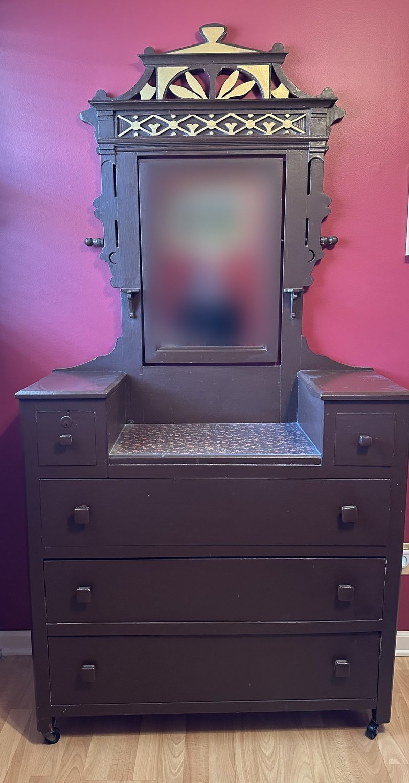 Victorian Eastlake-style dresser with carved fretwork crest, gold-painted geometric pediment, and full-length swing mirror — buried under thick brown paint, hiding all of the wood grain and most of the carved detail. Customer's reflection in the mirror has been blurred for privacy. Before restoration at Doll Furniture Company.