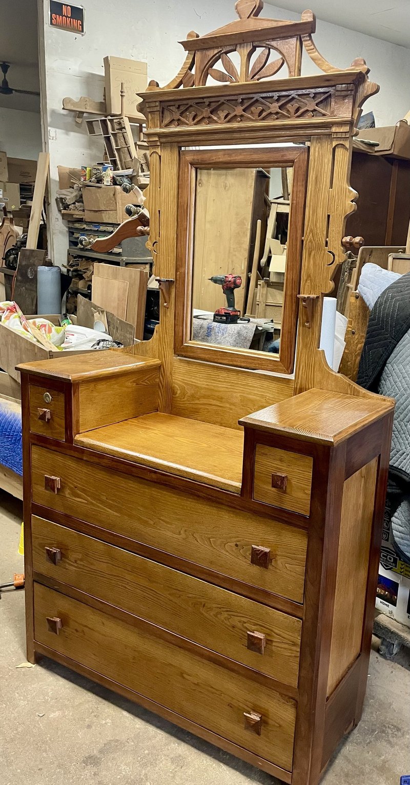 The same Victorian Eastlake dresser after stripping and restoration at Doll Furniture Company — the carved fretwork crest, scrolled mirror frame supports, fluted side standards, and beautifully figured ash drawer fronts are all fully revealed in their natural wood grain, with original square wooden drawer pulls preserved