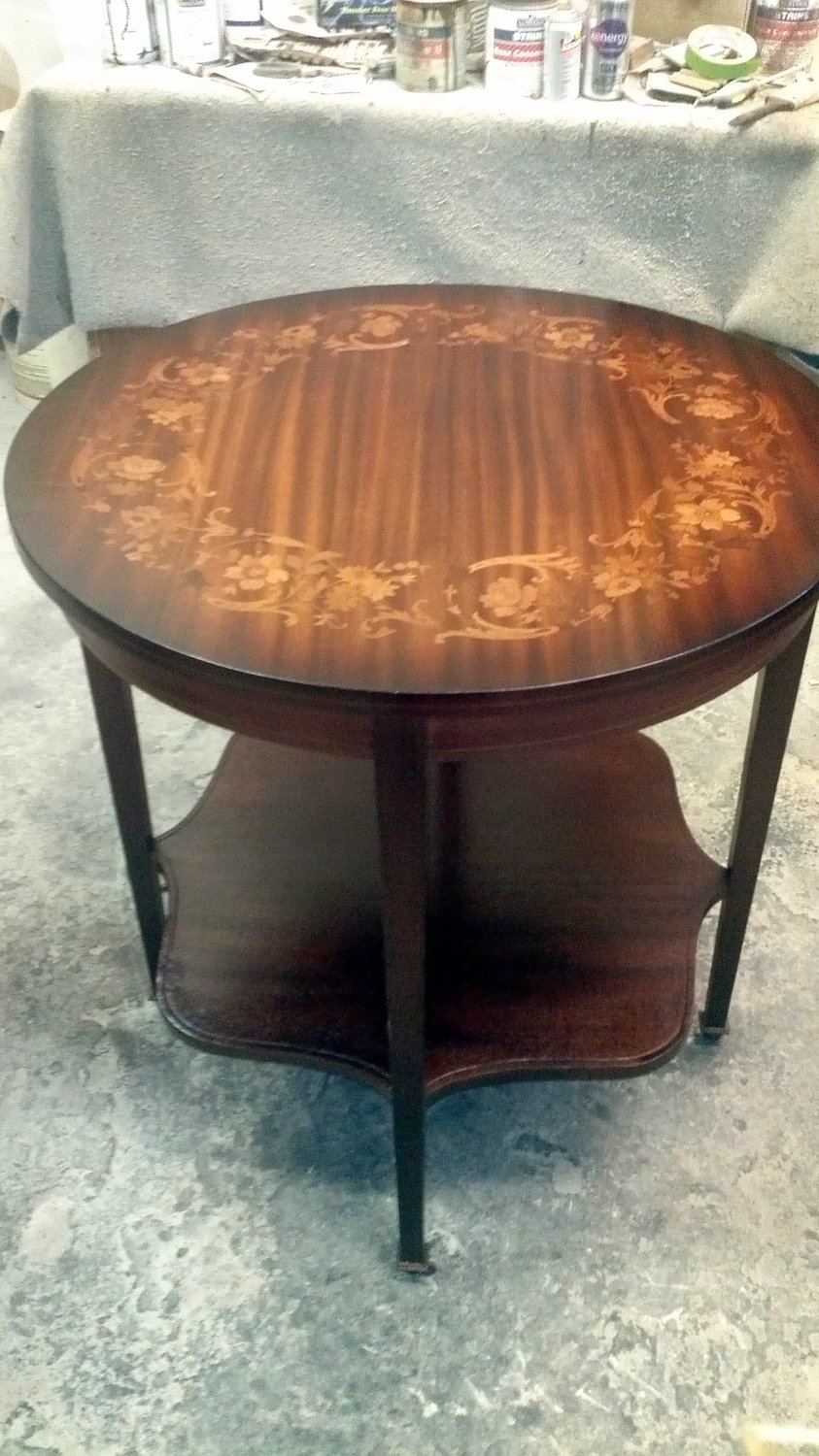 The same marquetry inlay parlor table after refinishing at Doll Furniture Company — deep mahogany stain brings out the floral inlay pattern in vivid contrast, satin top coat highlights the ribbon mahogany grain, and the lower scalloped shelf is restored to a rich dark finish