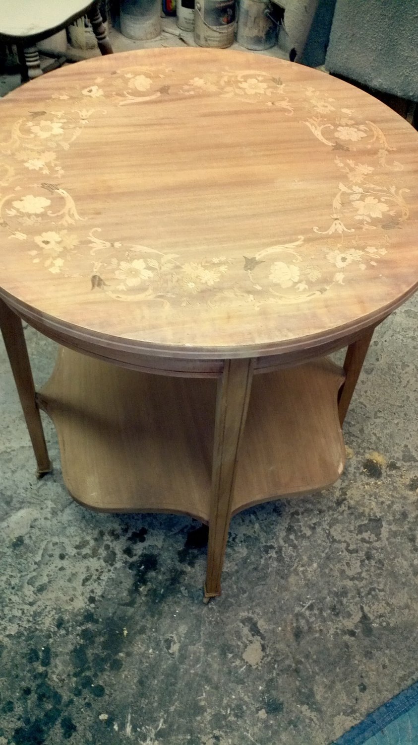 Antique round mahogany parlor table with intricate floral marquetry inlay along the perimeter, shown stripped to bare wood with faded, washed-out finish — before refinishing at Doll Furniture Company
