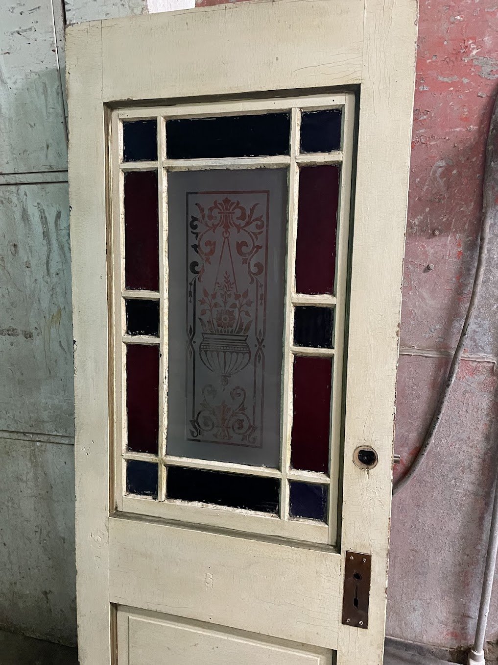 Antique Victorian Eastlake entry door covered in thick cream paint, hiding all wood grain and carved detail — stained glass border panels in red and dark blue still visible around the central etched glass panel with floral urn design — before restoration at Doll Furniture Company