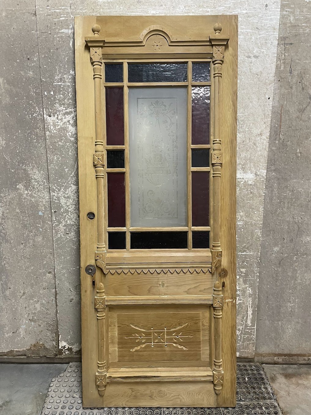 The same Victorian Eastlake entry door after chemical stripping in the Strip-o-lator at Doll Furniture Company — original solid pine wood grain fully revealed, with carved spindles, decorative panels, sawtooth trim, and the etched glass and stained glass border now showcased against bare wood